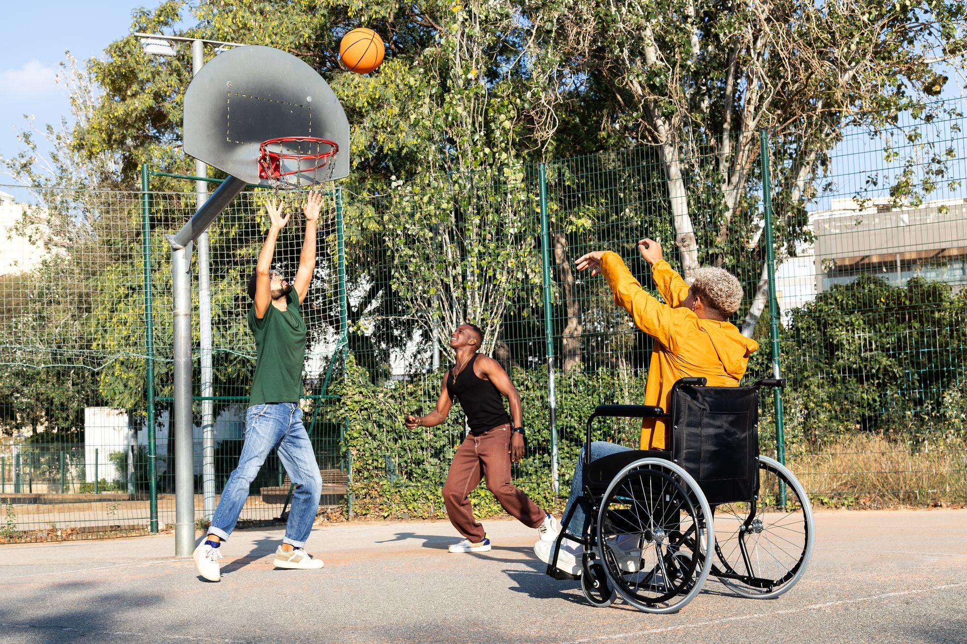 Tres personas jugando al baloncesto en una cancha al aire libre; una de ellas en silla de ruedas lanzando la pelota hacia el aro.