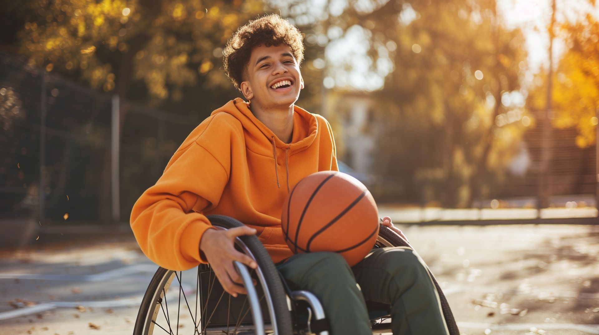 Adolescente sonriente en silla de ruedas sosteniendo una pelota de baloncesto en la cancha, vistiendo una sudadera con capucha naranja.
