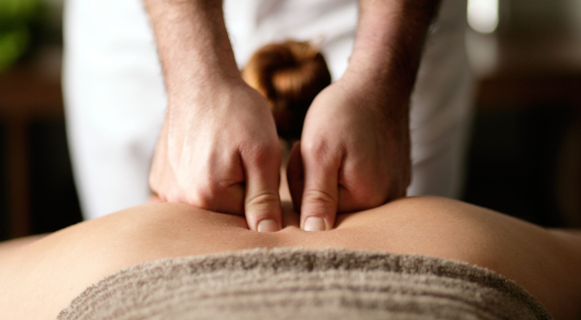 Hände massieren den Rücken einer Person. Die Masseurin übt mit den Fingerspitzen Druck auf den unteren Rücken aus.