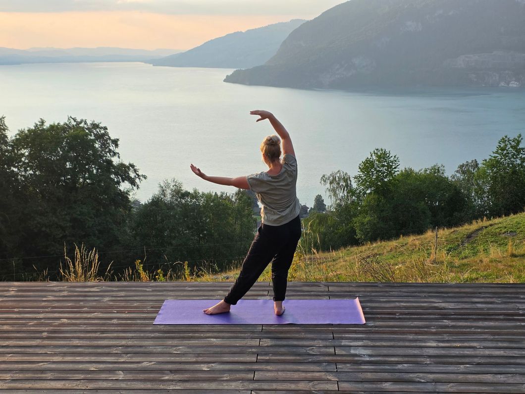 Eine Gruppe macht Yoga mit erhobenen Armen im Freien mit Blick auf einen See und die Berge.