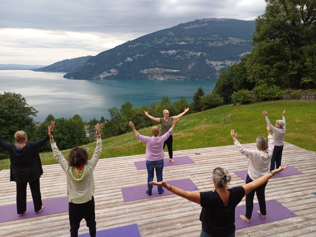 Eine Gruppe macht Yoga mit erhobenen Armen im Freien mit Blick auf einen See und die Berge.