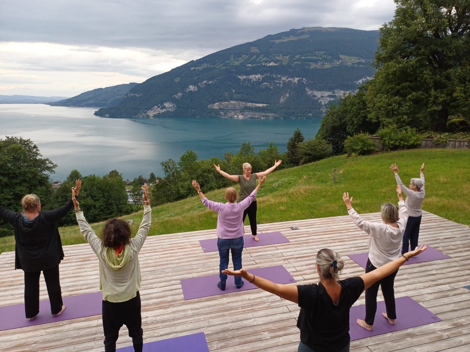 Eine Gruppe von Menschen macht Yoga mit erhobenen Armen und blickt auf einen See und Berge.