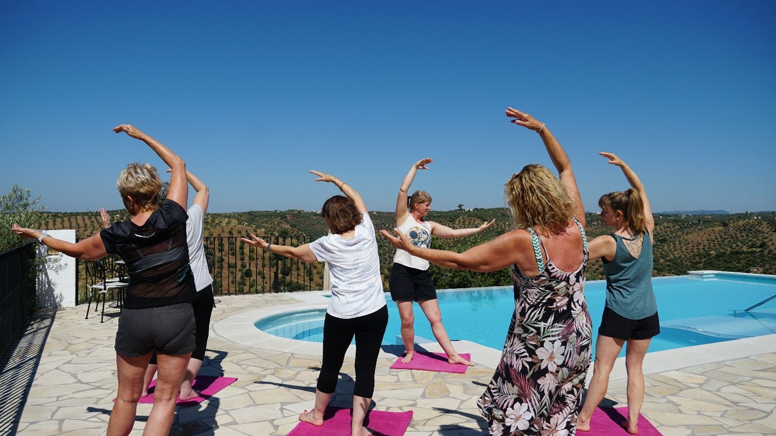 Eine Gruppe von Frauen macht an einem sonnigen Tag im Freien neben einem Pool Yoga, die Arme nach oben gewölbt.