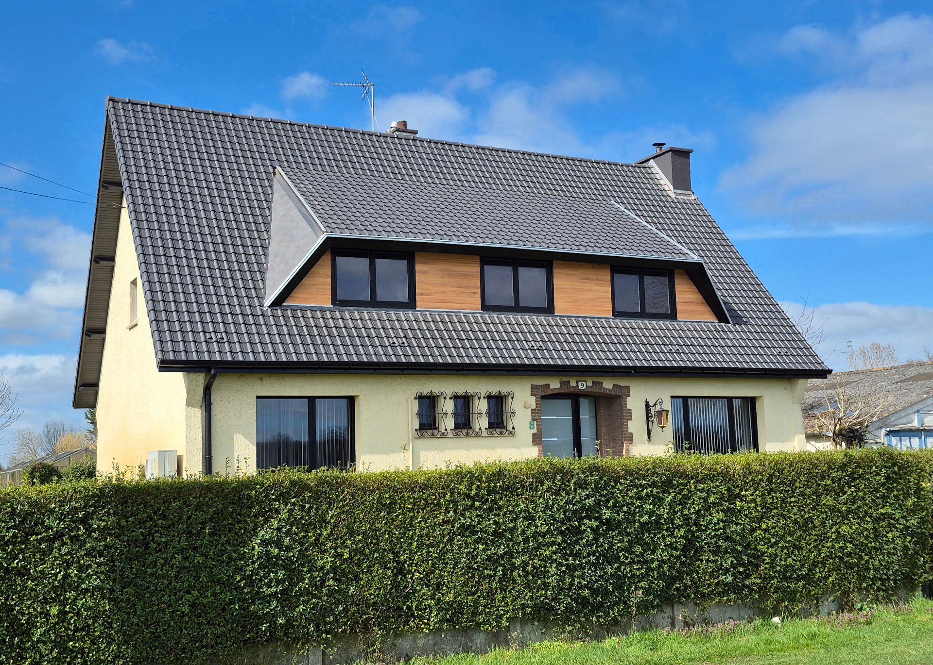 Maison en briques au toit de tuiles rouges, avec des échafaudages et une voiture garée devant. Ciel couvert.