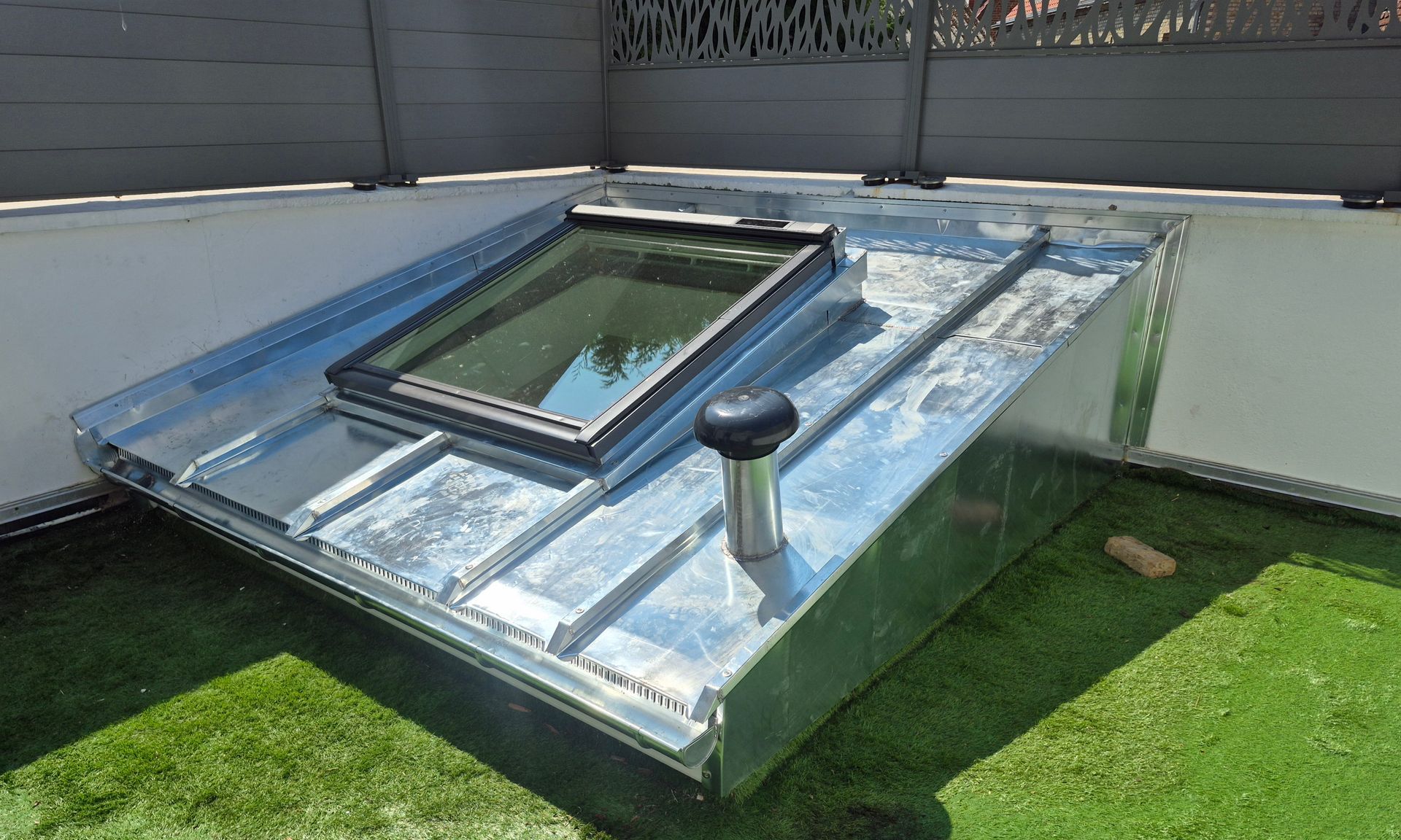 Puits de lumière métallique sur un toit, avec un tuyau de ventilation noir, installé sur du gazon artificiel vert.