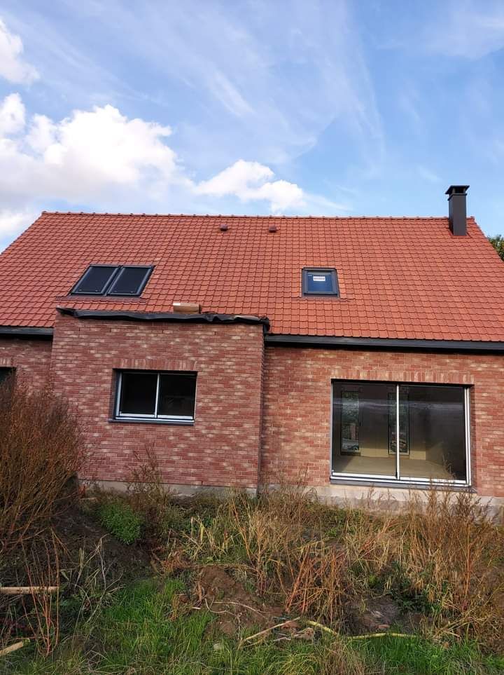 Maison en briques rouges avec un toit de tuiles rouges, cheminée noire, fenêtres et panneaux solaires sur fond de ciel bleu.