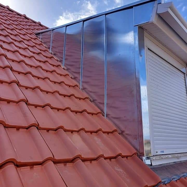 Toit en tuiles rouges avec des solins métalliques et une fenêtre avec un volet blanc fermé.