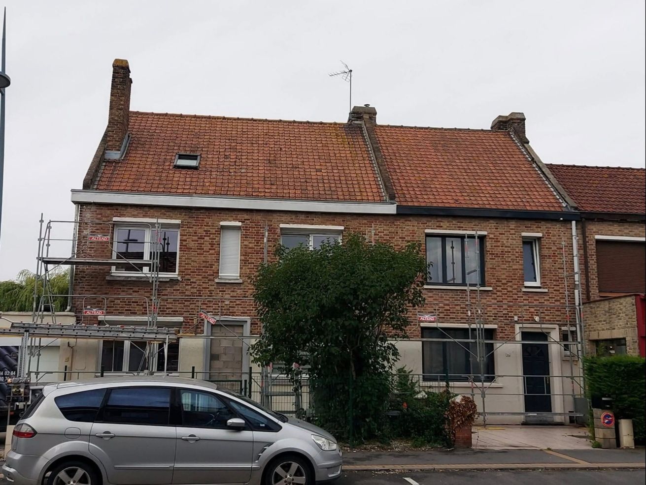 Maison en briques au toit de tuiles rouges, avec des échafaudages et une voiture garée devant. Ciel couvert.
