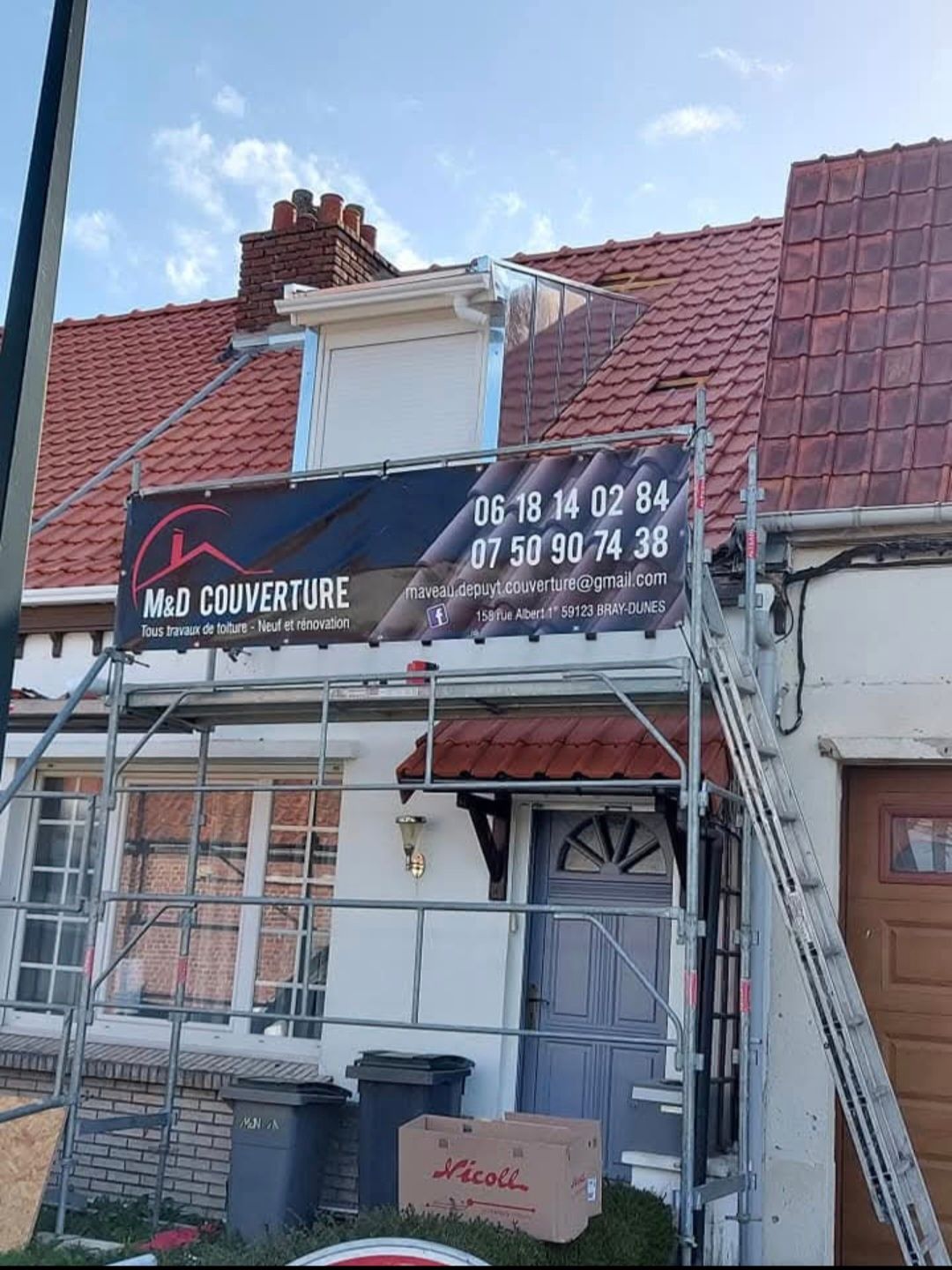 Échafaudage installé près d'une maison blanche dont le toit de tuiles rouges a été partiellement remplacé ; panneau publicitaire visible.