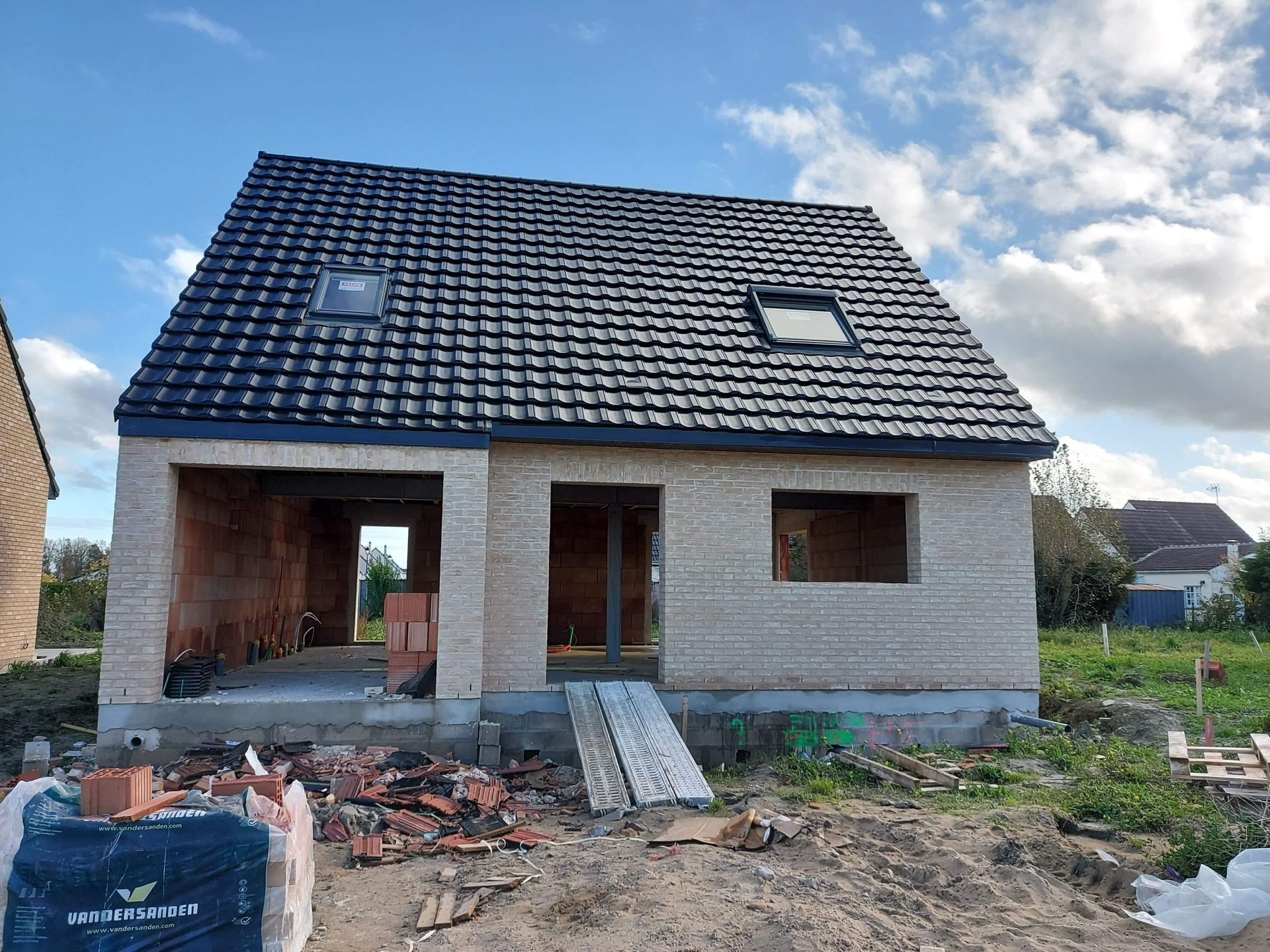 Une maison en briques en construction, avec un toit de tuiles sombres, une porte ouverte et deux puits de lumière se détachant sur un ciel bleu.