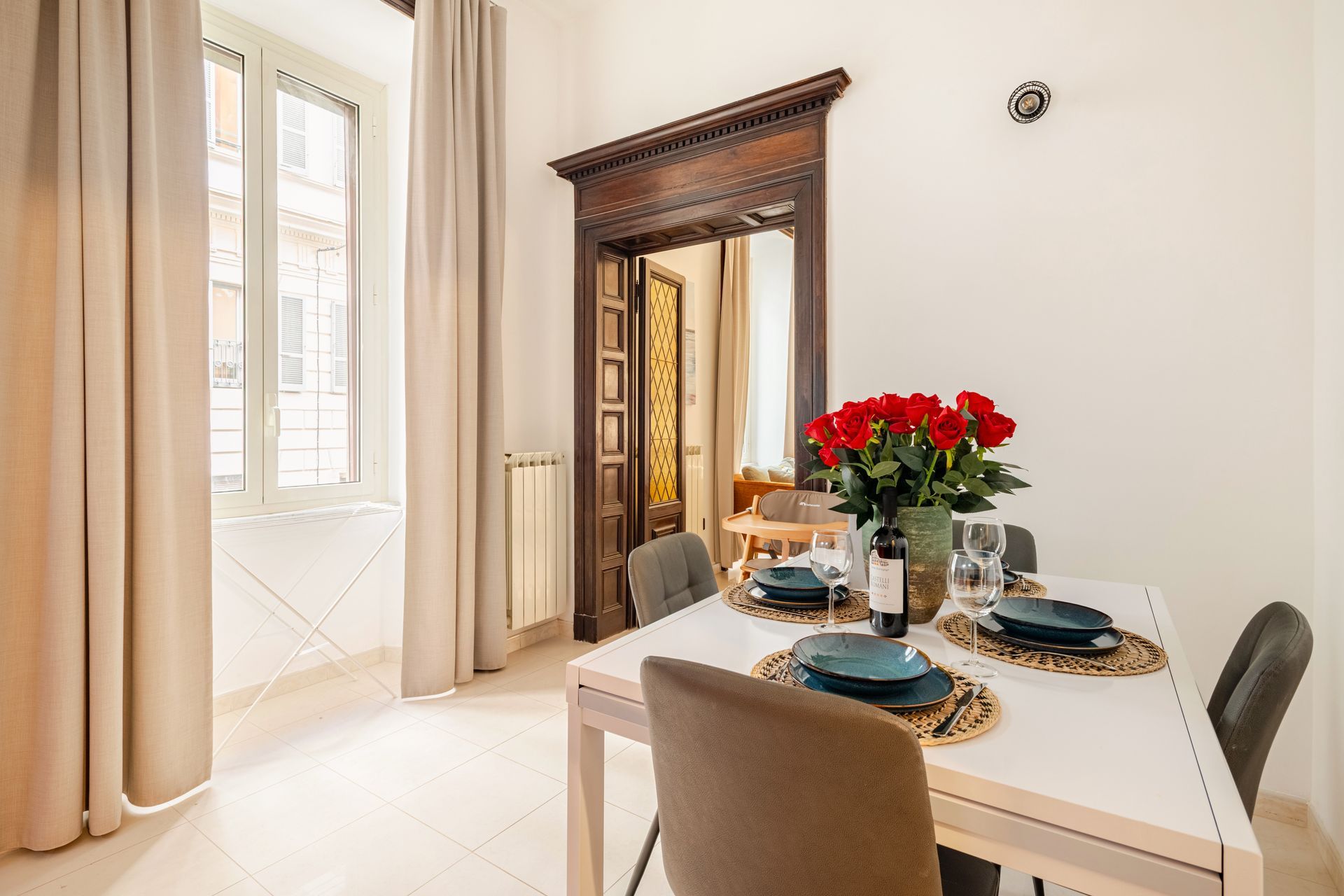 Dining area with table set for four, red roses, ornate doorway, window with beige curtains.