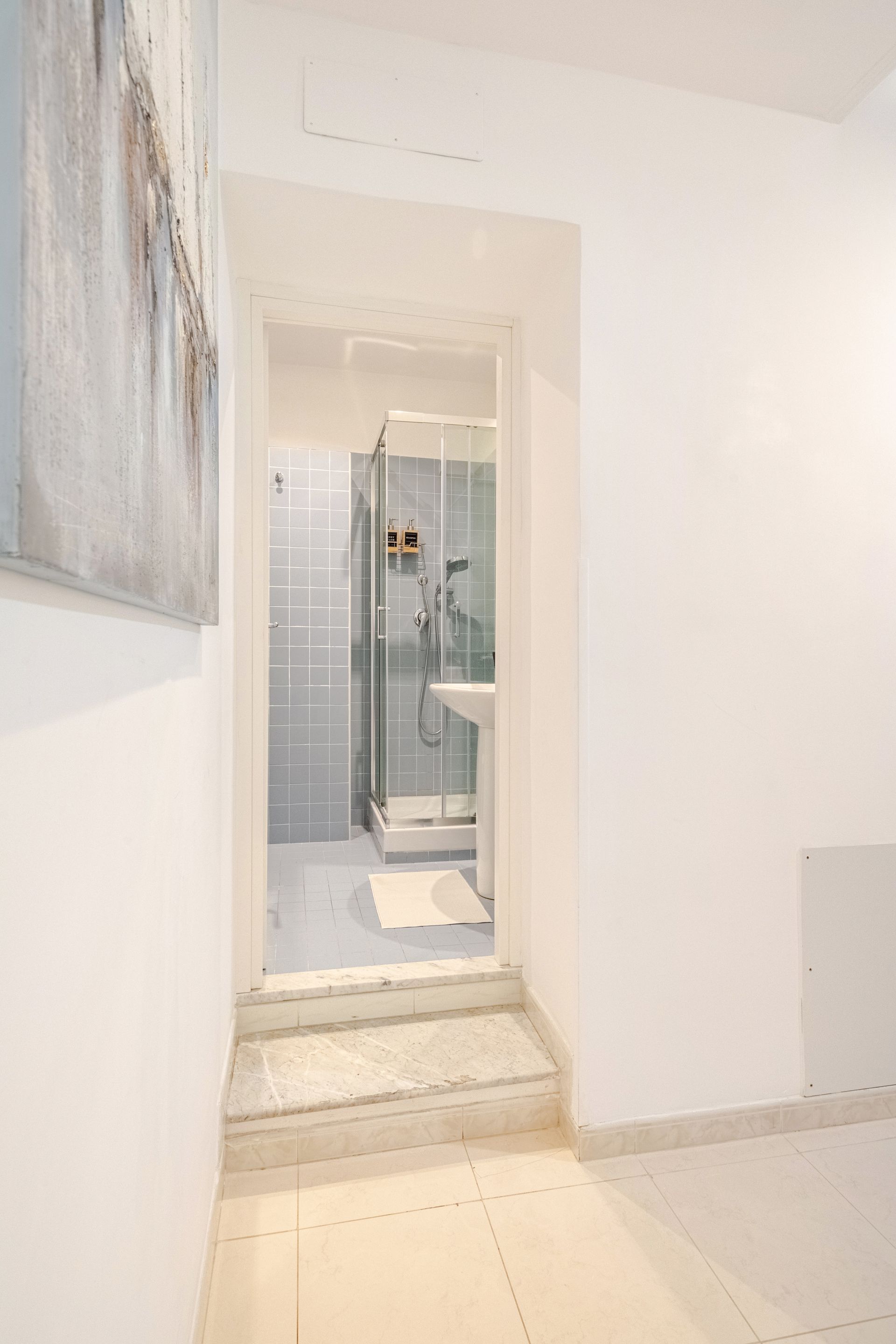 Narrow white hallway leading to a bathroom with a shower. Light gray tile walls.