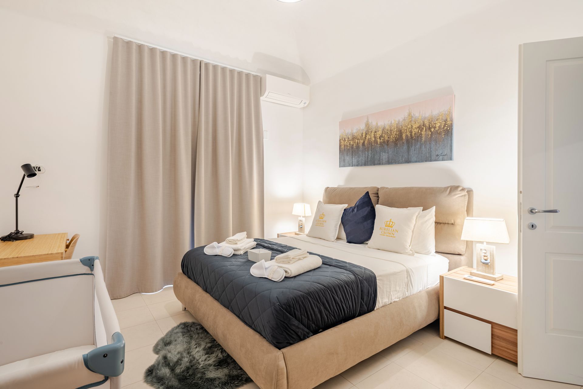 Bedroom with a bed, crib, and neutral decor; includes a small desk, art, and lamps.