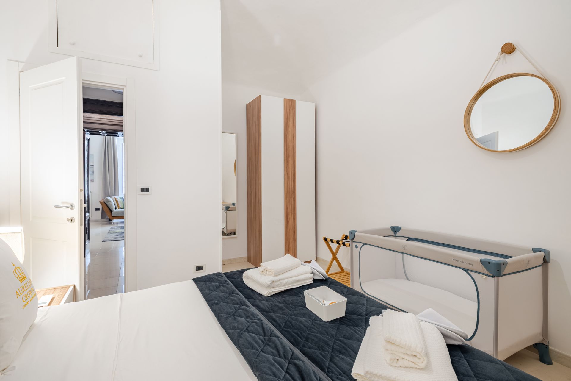 Bedroom with white walls, bed with navy blanket, crib, wardrobe, mirror, towels.