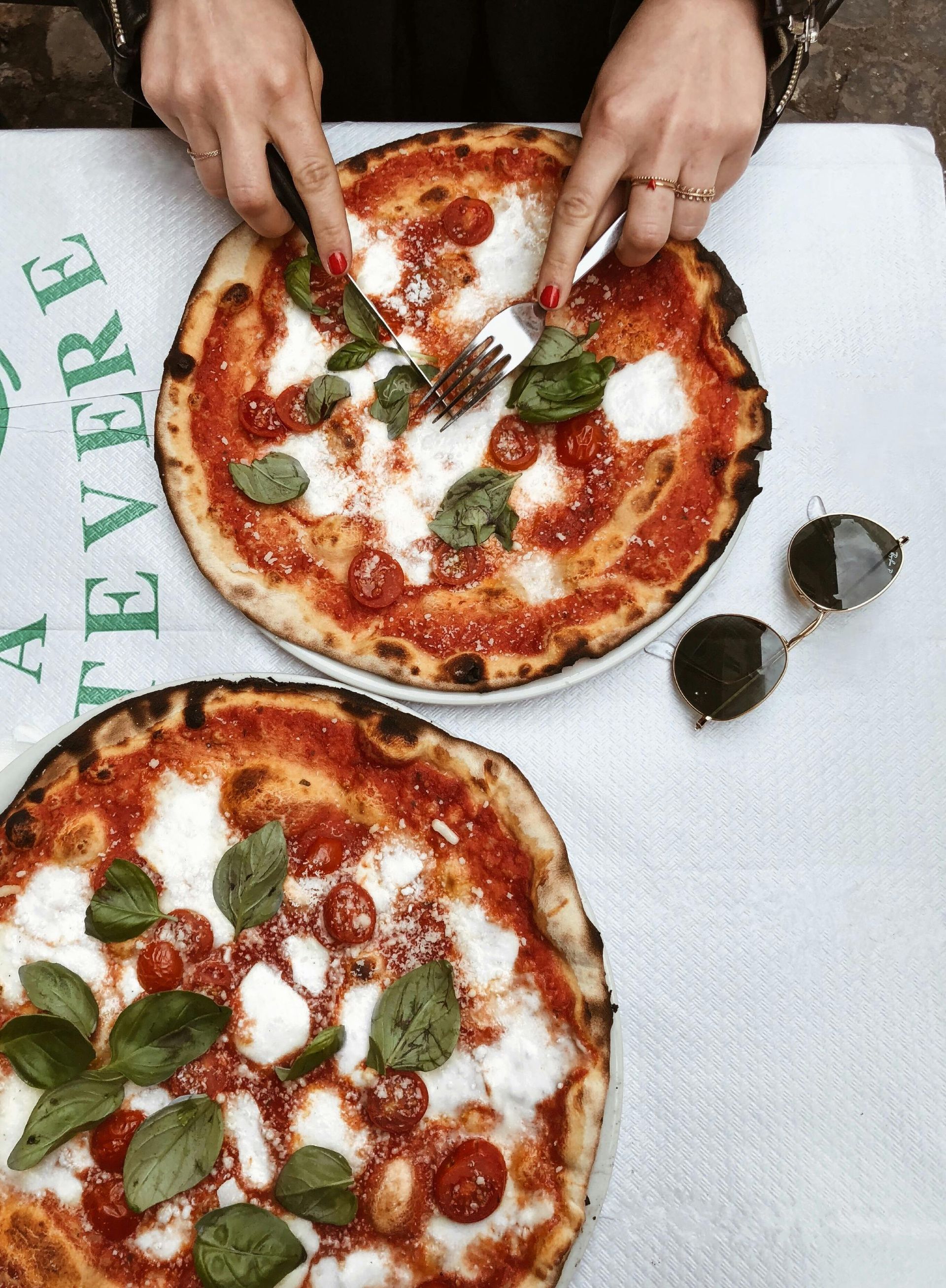 Two margherita pizzas on a white tablecloth, with hands cutting one and sunglasses beside it.