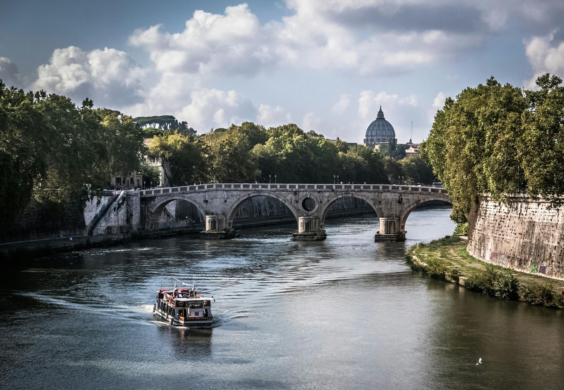 A boat on the Tiber River in Rome, passing under a stone bridge with the dome of St. Peter's Basilica in the distance.
