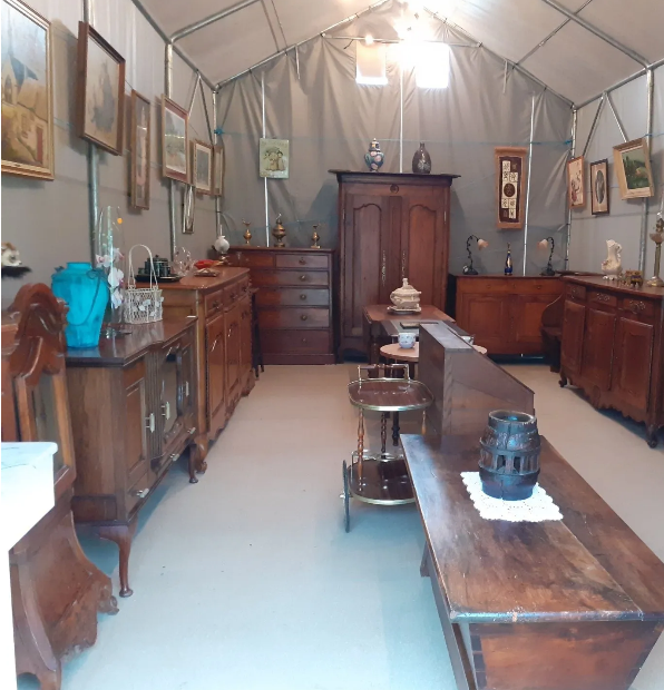À l'intérieur d'une tente : exposition de meubles anciens, notamment des armoires en bois, des tables et des tableaux encadrés.