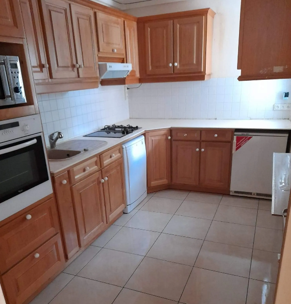 Une petite cuisine avec des armoires marron clair, des appareils électroménagers blancs et un sol carrelé.