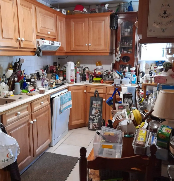 Cuisine encombrée avec des armoires en bois clair, pleine d'objets sur les comptoirs, la table et les étagères.