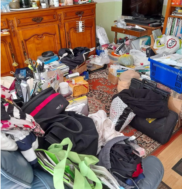 Salon en désordre : vêtements, livres et objets divers éparpillés sur le sol et les meubles.