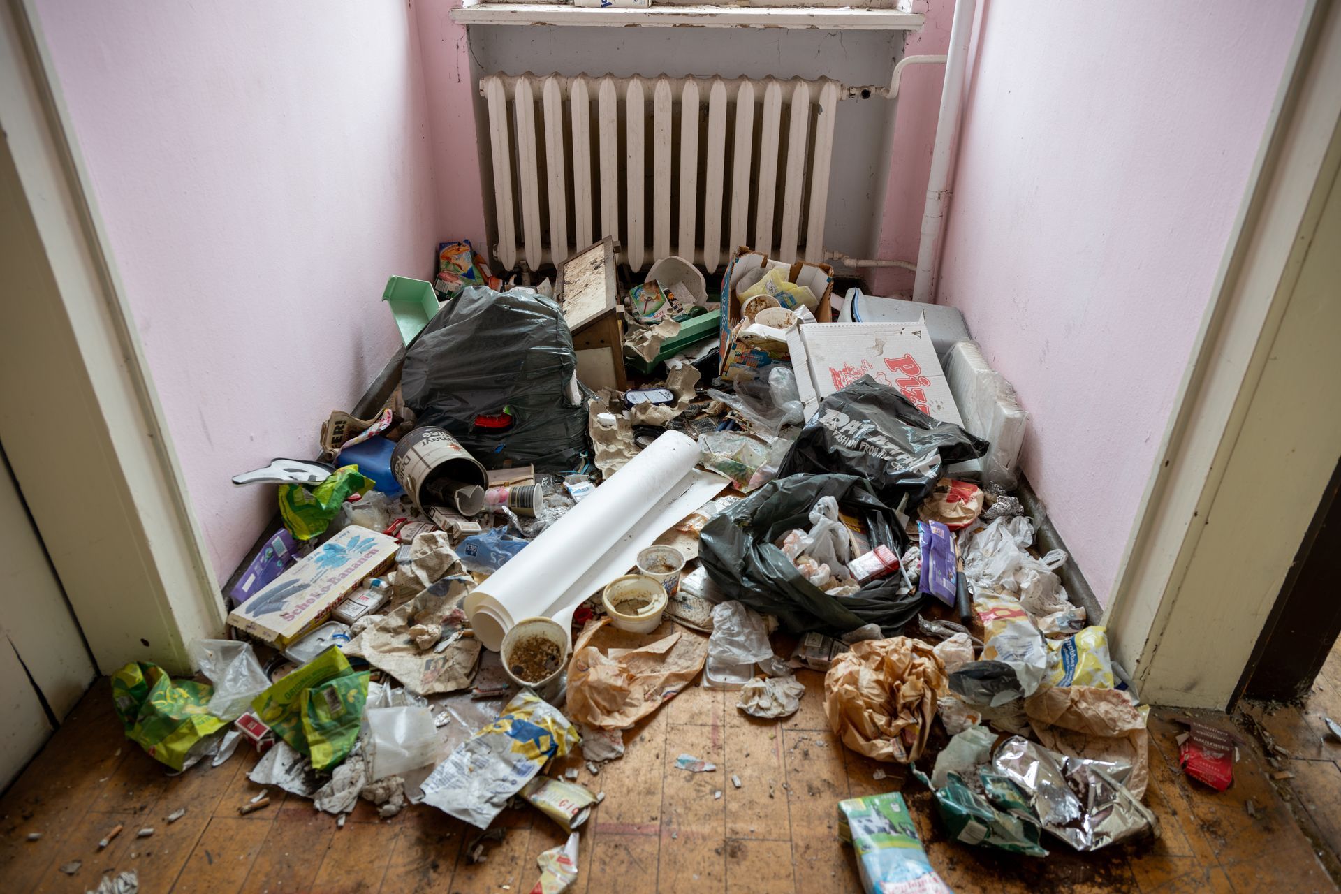Une petite pièce remplie de déchets éparpillés, un radiateur contre le mur, des murs roses et un plancher en bois.