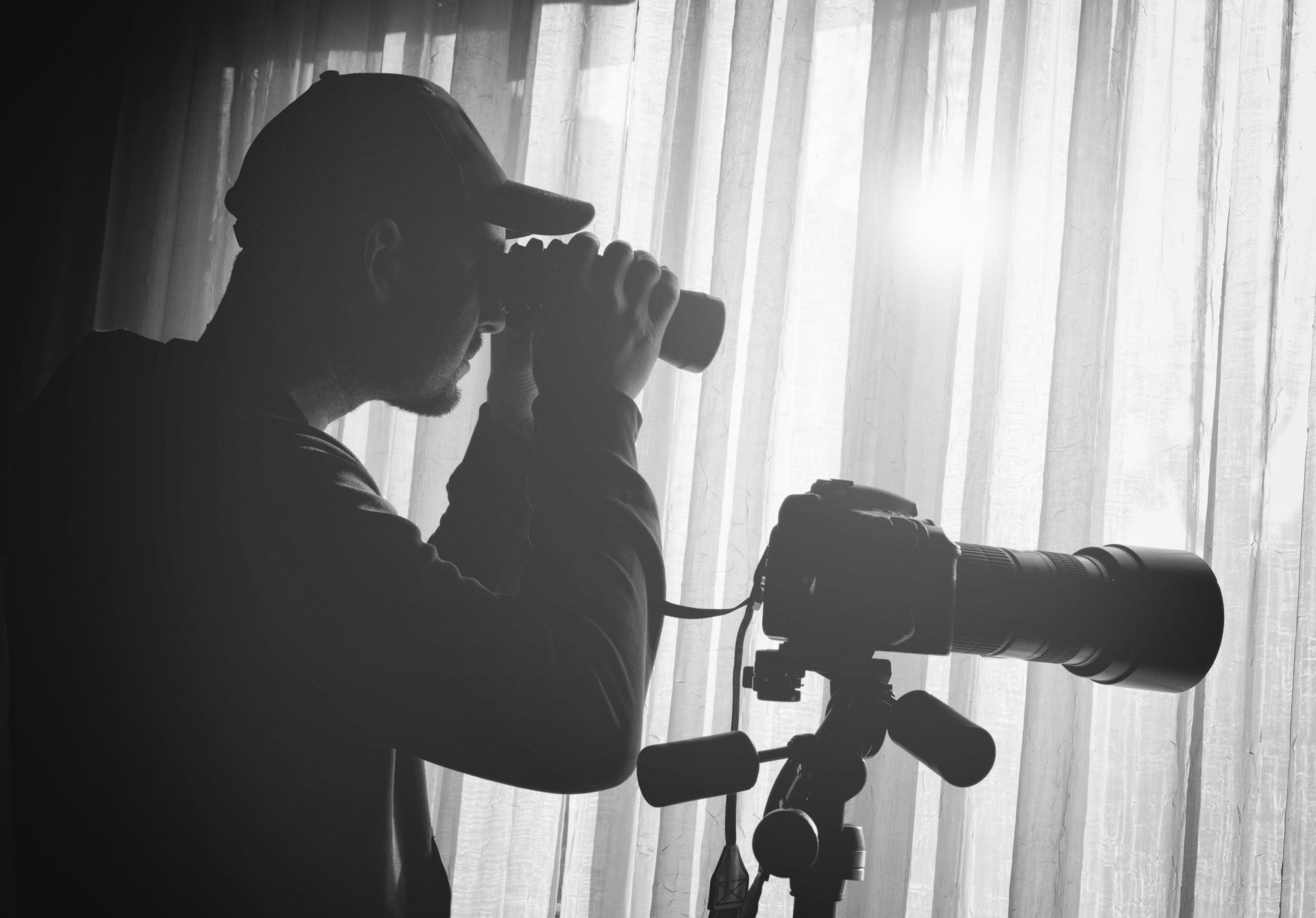 Silhouette d'un photographe regardant à travers un appareil photo, à côté d'un téléobjectif monté sur un trépied, derrière des rideaux transparents.