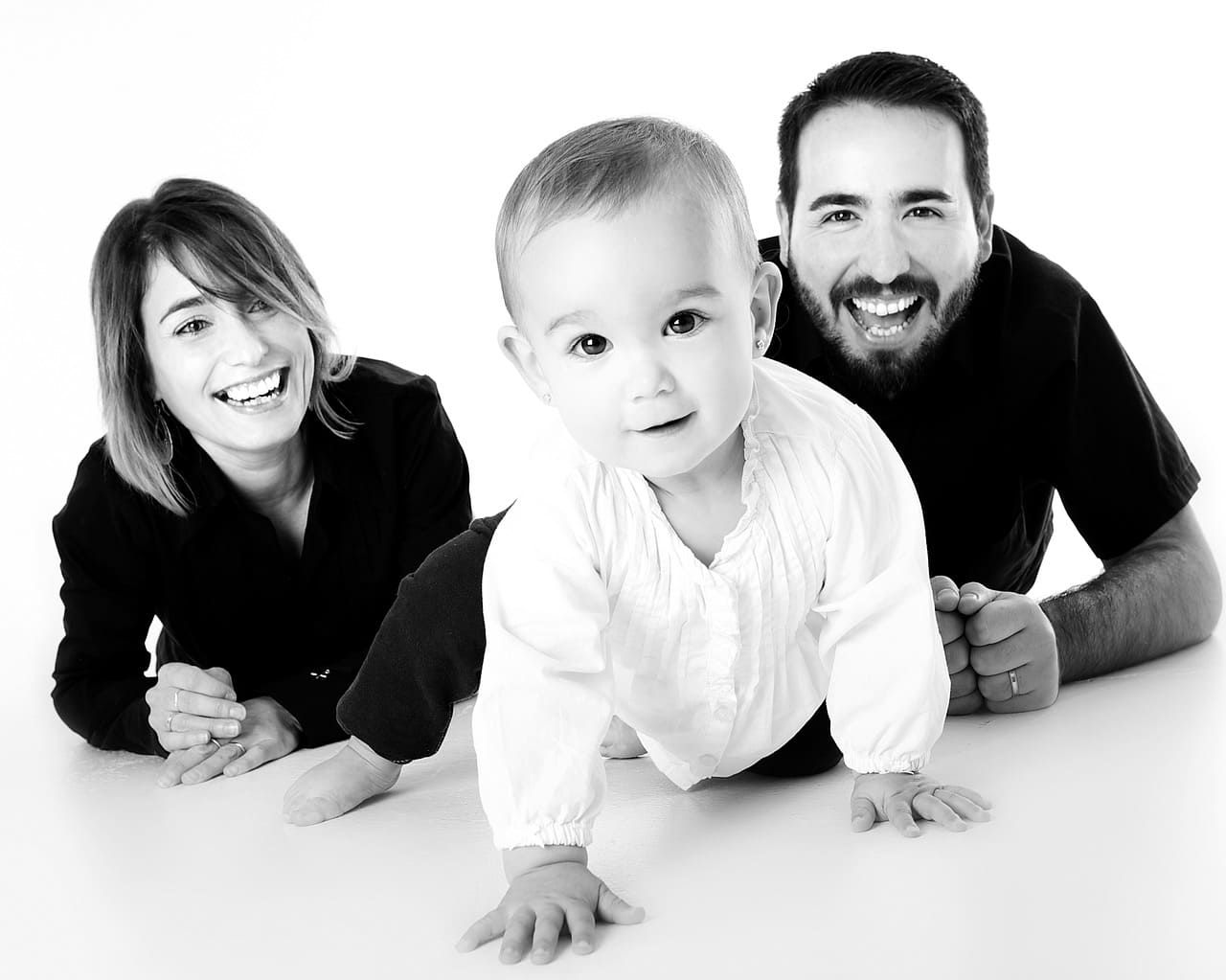 Une famille de trois personnes souriantes, allongées sur un sol blanc, avec un bébé devant et deux adultes derrière.