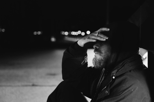 Photo en noir et blanc d'une personne assise la nuit, la main sur le visage, dans une rue faiblement éclairée.