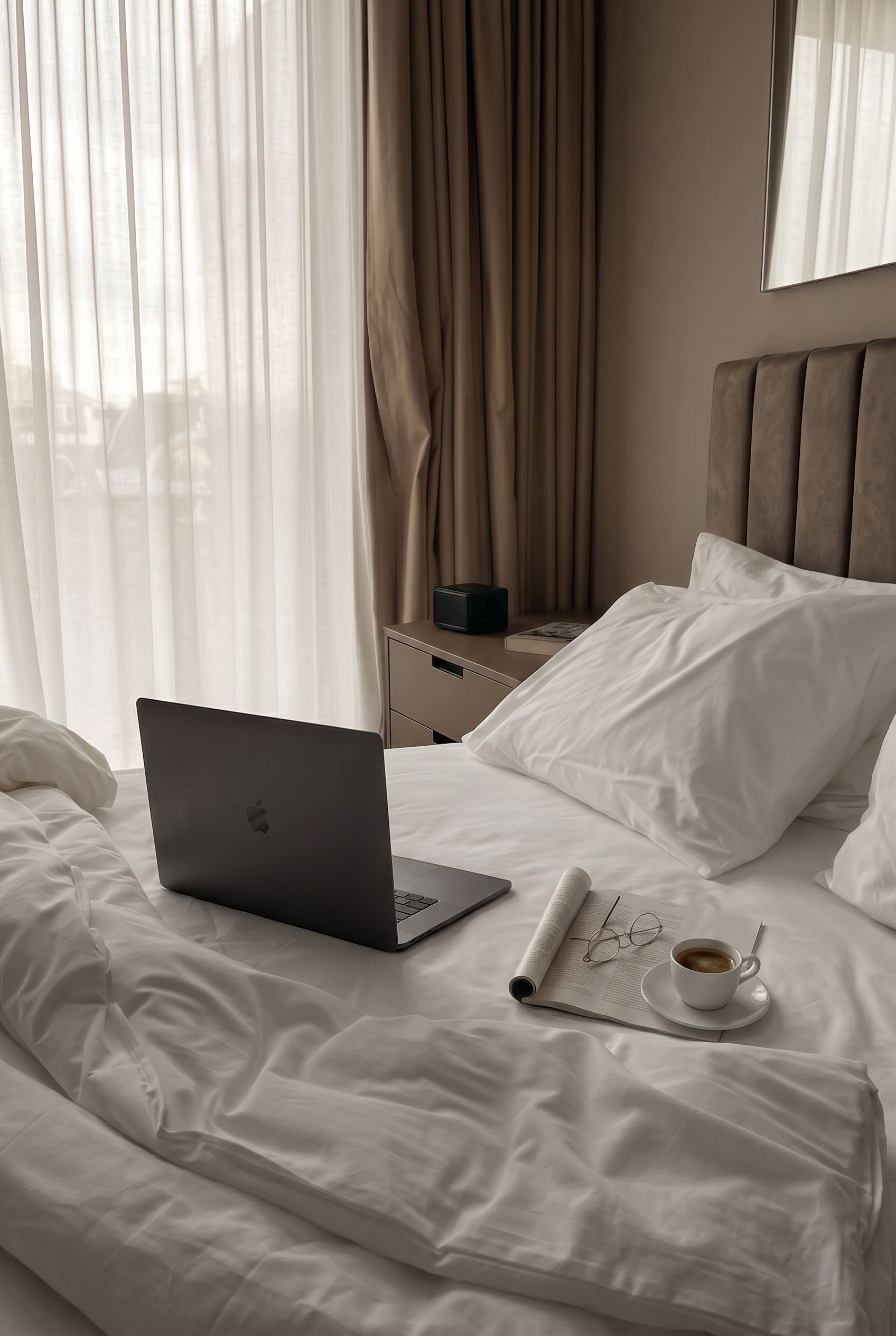 Laptop auf einem zerwühlten weißen Bett in einem hellen Schlafzimmer, neben einem Tablett mit Kaffee und einer Blumenvase