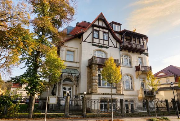 Historische weiße Villa und kunstvollen Balkonen hinter einem schwarzen Zaun, eingerahmt von herbstlichen Bäumen