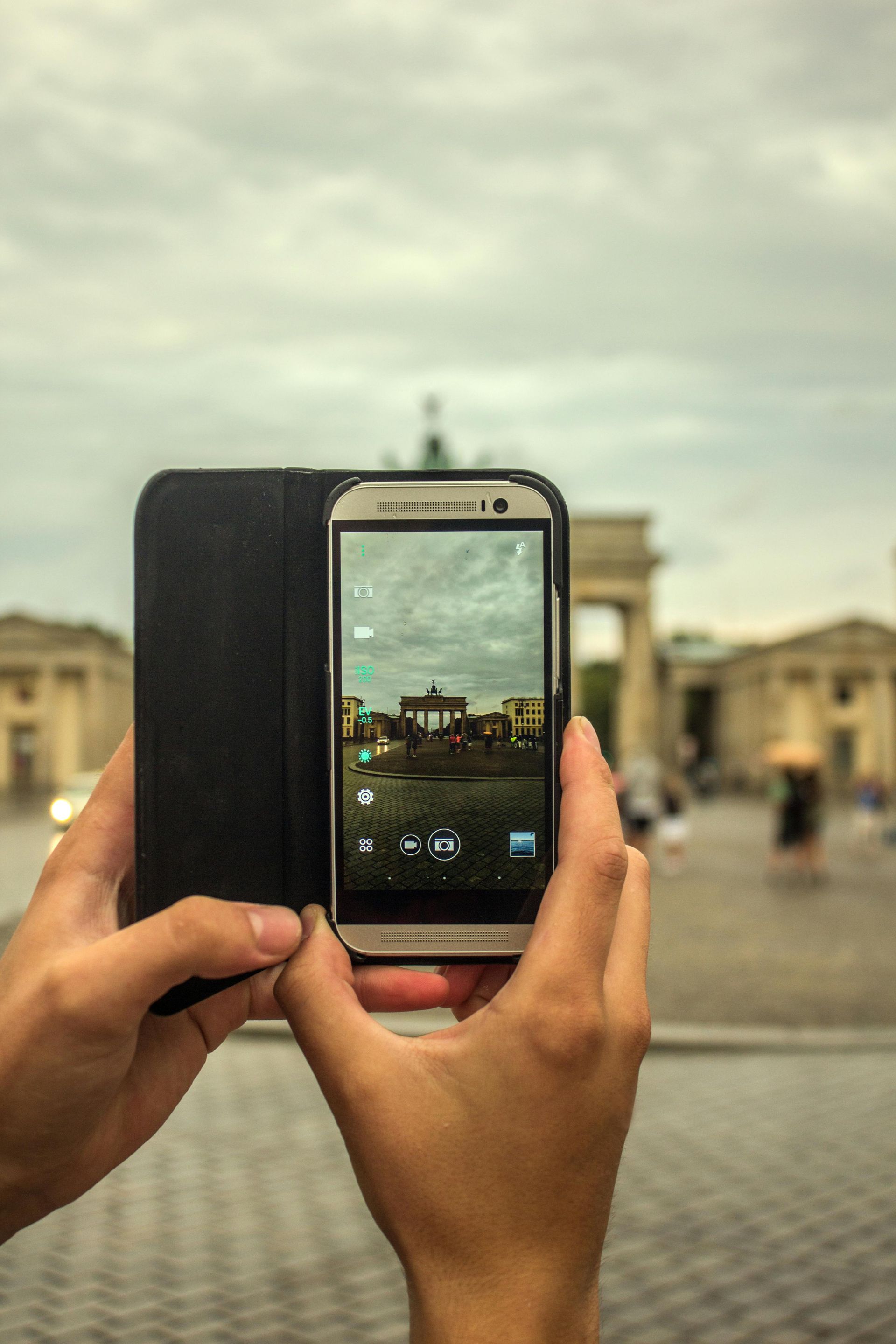 Hände halten ein Smartphone und fotografieren einen Platz mit Gebäuden im Hintergrund.