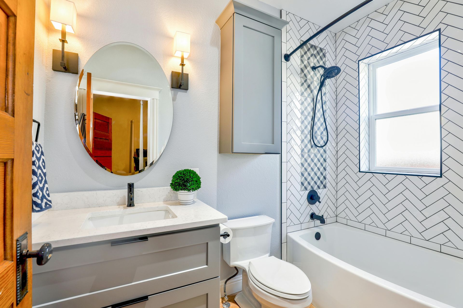 Helles Badezimmer mit Waschtisch, rundem Spiegel, Toilette und weißer Badewanne mit gemustertem Duschvorhang und Fliesen.
