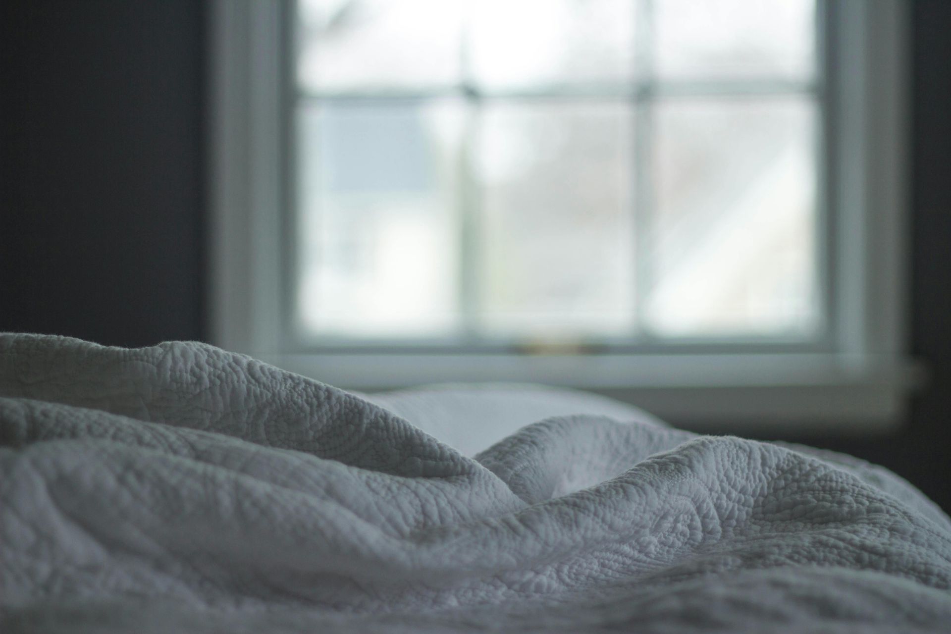 Graue Decke auf einem Bett neben einem hellen Fenster in einem sanft beleuchteten Raum