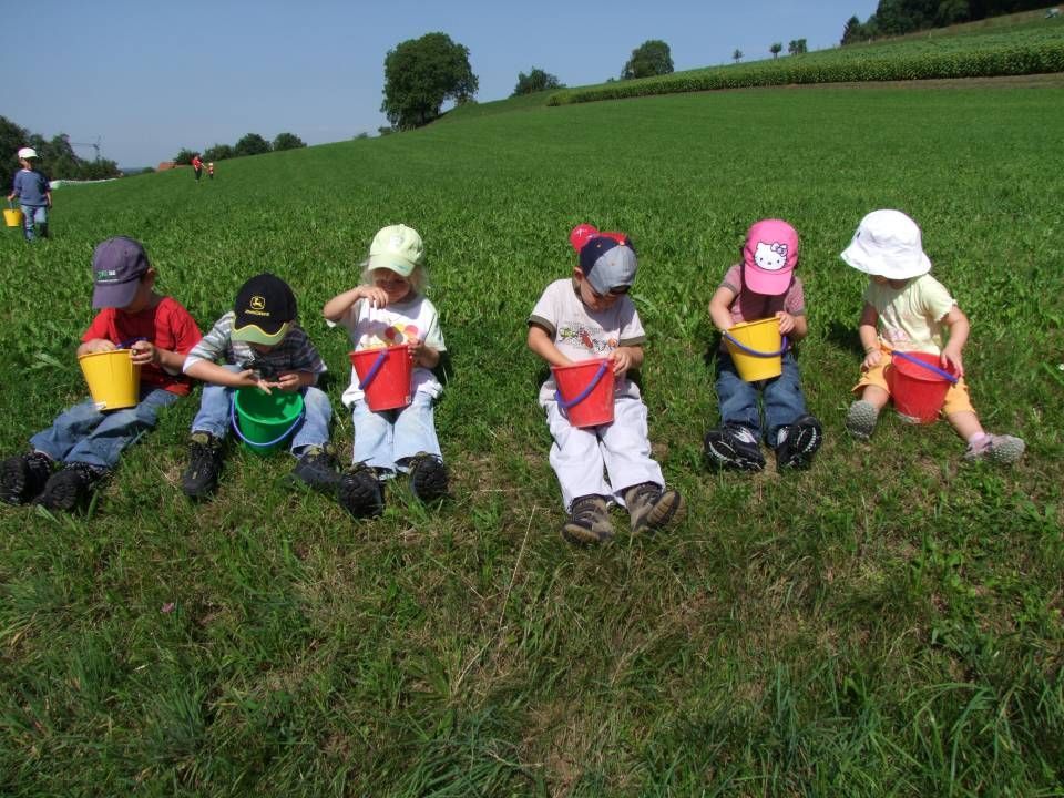 Die Kinder sitzen in einer Reihe auf einem grasbewachsenen Hügel, jedes hält einen bunten Plastikeimer.