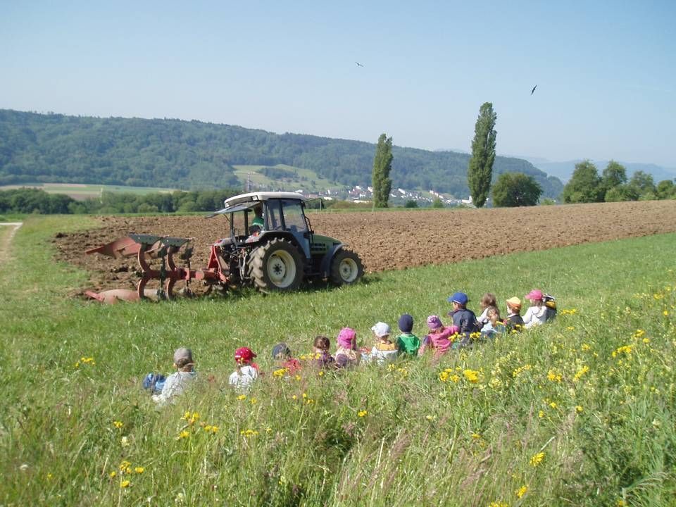 Ein Traktor pflügt ein Feld, während eine Gruppe Kinder in der Nähe im Gras sitzt und zuschaut; im Hintergrund erheben sich Berge.