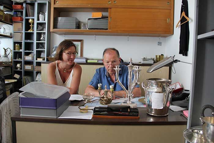 Une femme et un homme sont assis à un bureau avec des objets en argent, probablement dans un magasin ou un bureau.