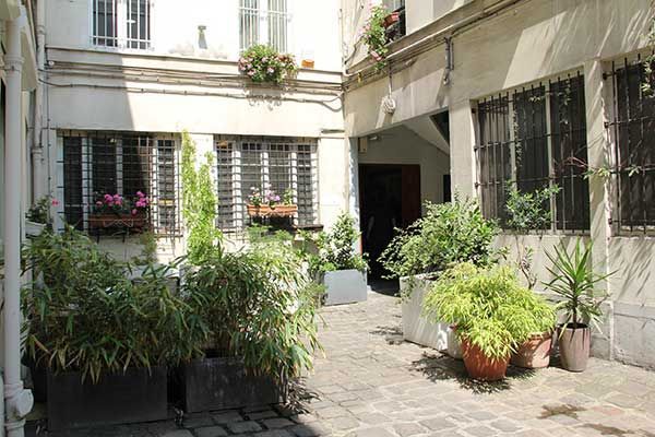 Une cour ensoleillée avec des plantes en pots et des jardinières, un pavage en pierre et des bâtiments blancs.