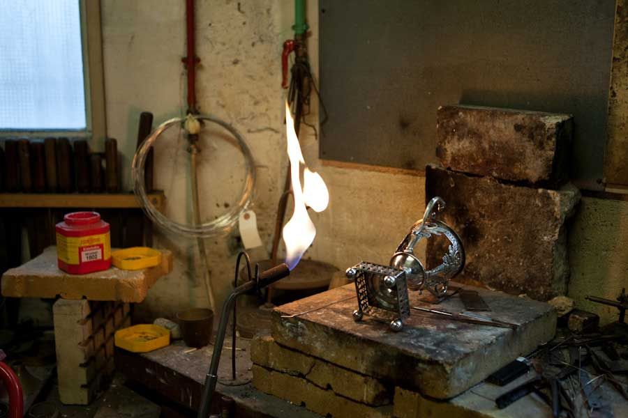 Une sculpture en métal est chauffée au chalumeau dans un atelier. On aperçoit des flammes, des briques et des outils.