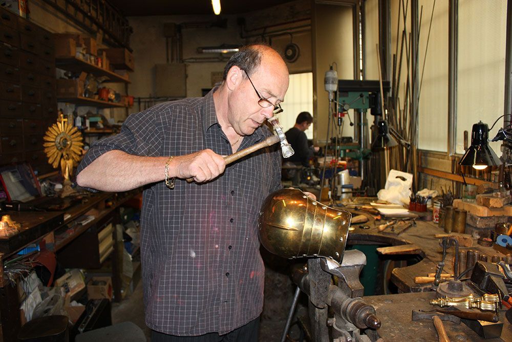 Un homme martèle un objet en laiton dans un atelier. Une autre personne travaille à l'arrière-plan.