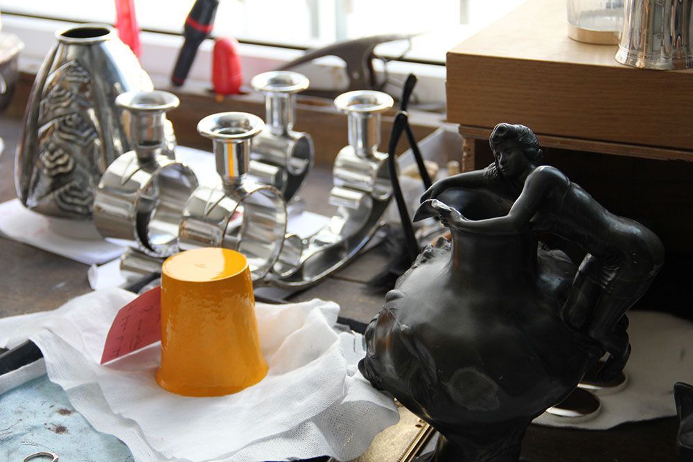 Gros plan sur des objets en argent et un vase sculpté sombre posés sur un plan de travail, une tasse jaune sur une nappe blanche.