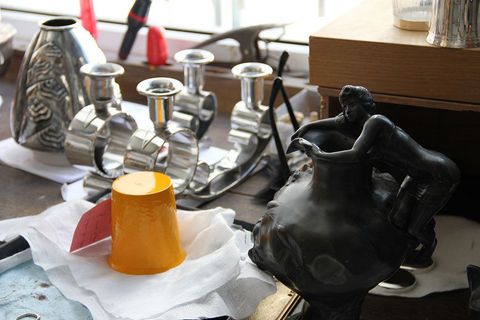 Gros plan sur des objets en argent et un vase sculpté sombre posés sur un plan de travail, une tasse jaune sur une nappe blanche.