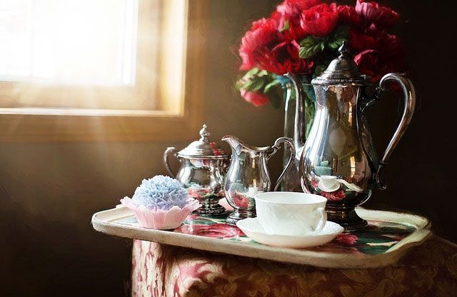 Un service à thé en argent, des roses et un cupcake sur un plateau près d'une fenêtre ensoleillée.