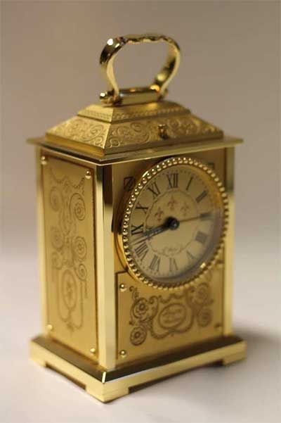 Pendule de voyage dorée avec chiffres romains, poignée et gravures florales.