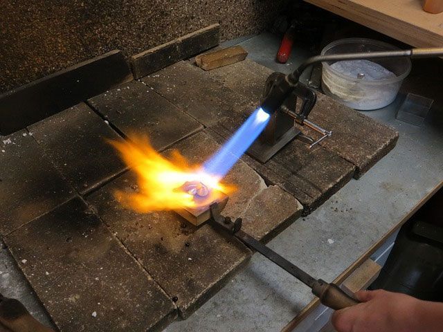 Un bijoutier brûle du métal au chalumeau sur une surface en briques réfractaires, avec des flammes bleues et oranges.