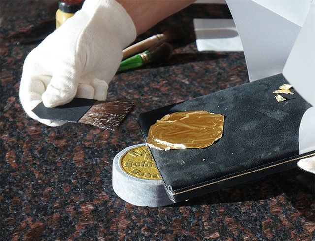 Personne portant des gants blancs manipulant des feuilles d'or au pinceau sur une surface noire.