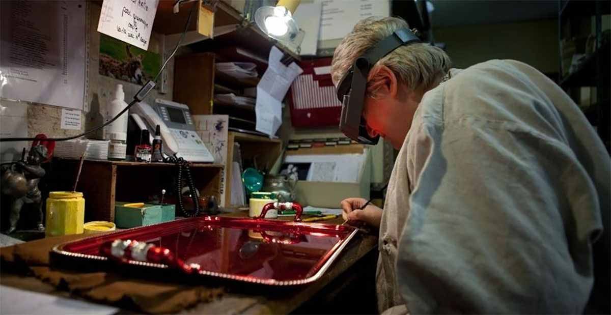 Une personne portant une visière grossissante travaille sur un plateau rouge orné d'un motif détaillé et complexe dans un atelier encombré.
