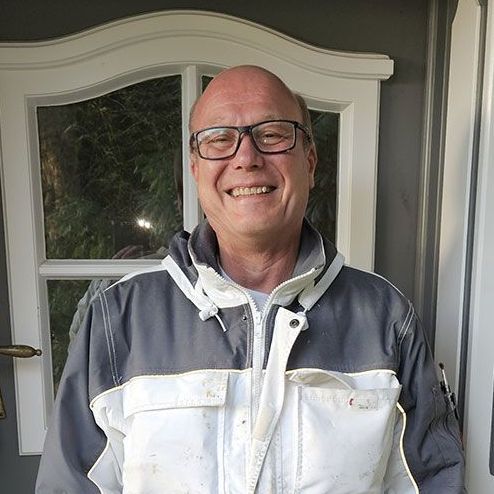 Ein Mann mit Brille und Jacke steht lächelnd vor einer Tür.