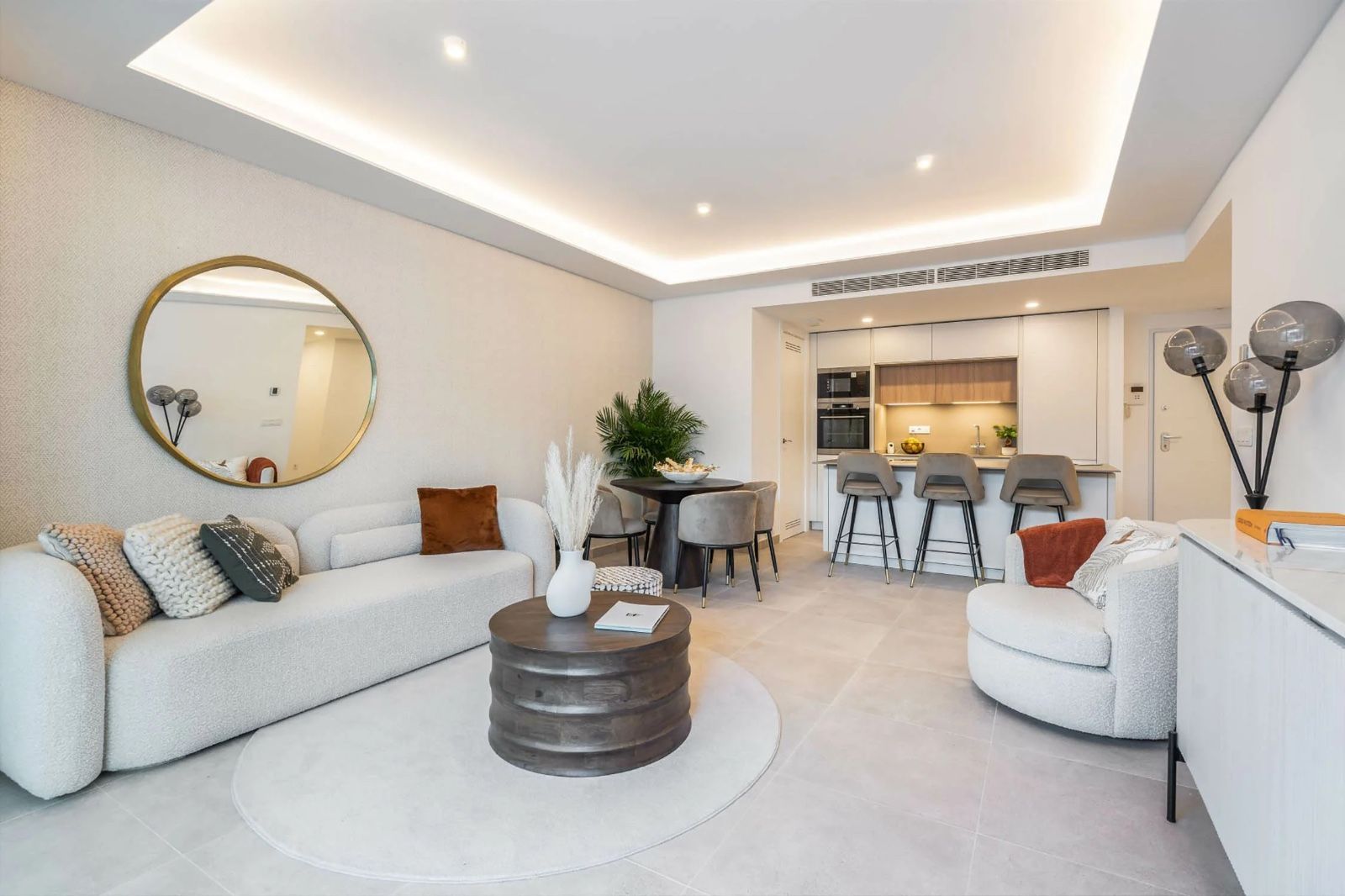 Luminoso salón moderno con sofás blancos, espejo redondo, mesa de centro y cocina abierta con taburetes de bar.