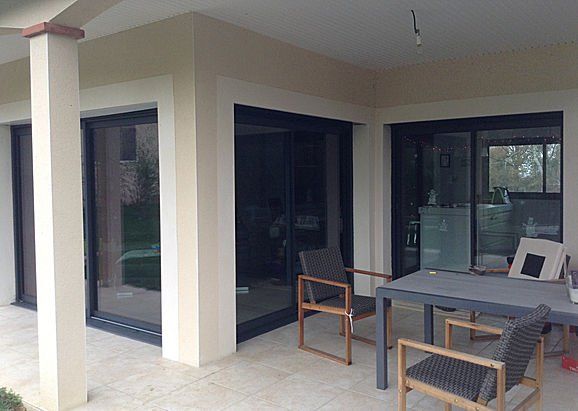 Patio avec portes coulissantes en verre à cadre foncé, chaises, table et sol en pierre, avec un extérieur de couleur crème.