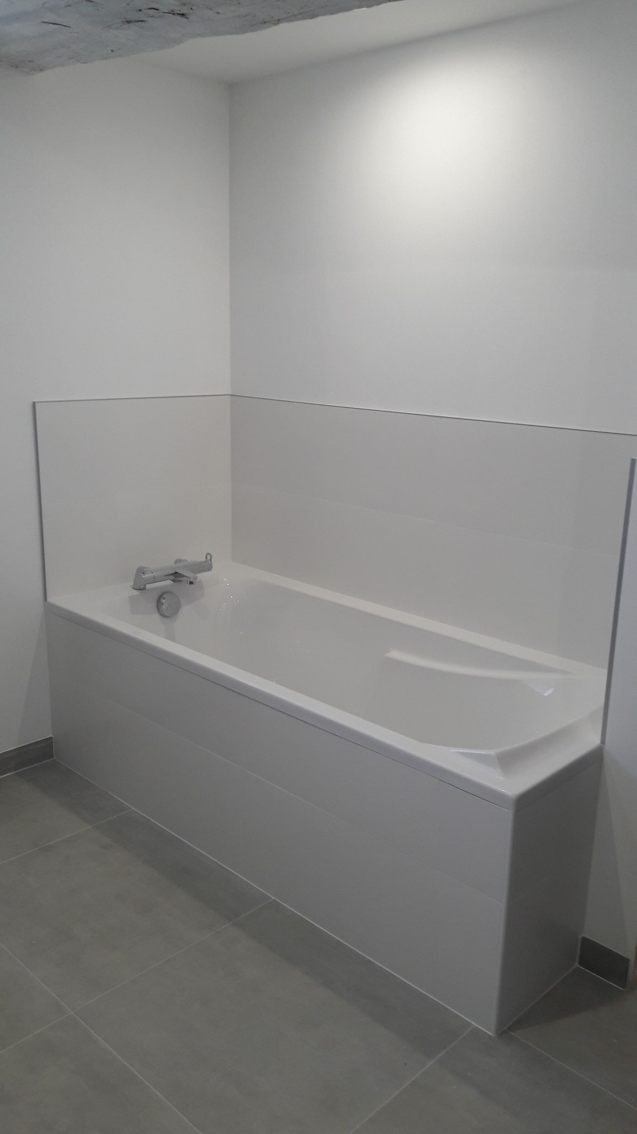 Baignoire avec paroi et robinet blancs dans une pièce au sol gris clair.